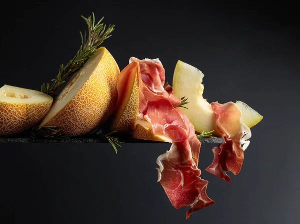 Prosciutto med melon och rosmarin på svart bakgrund. — Stockfoto