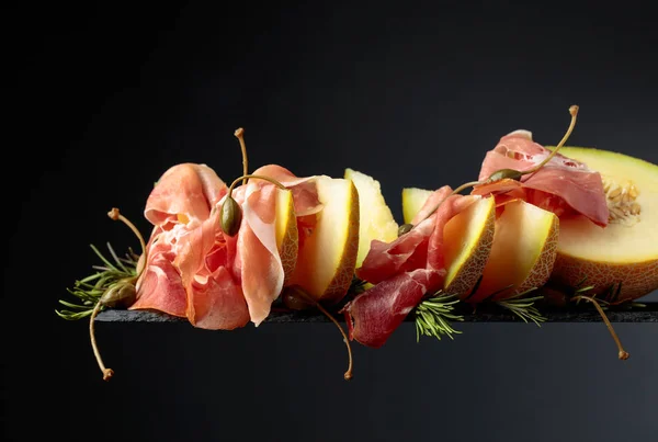 Schinken mit Melone, Rosmarin und Kapern auf schwarzem Hintergrund — Stockfoto