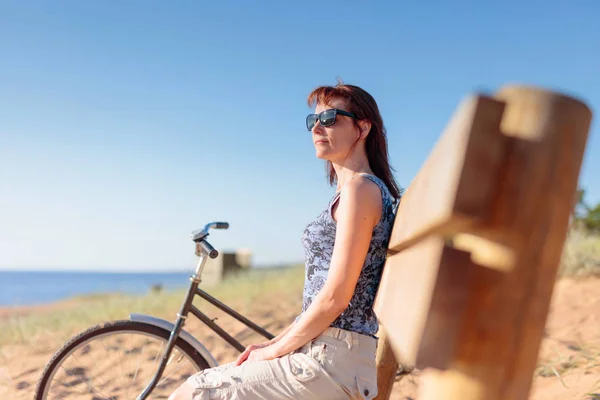 Mulher de meia-idade veio em uma bicicleta para a praia e descansar sentar — Fotografia de Stock