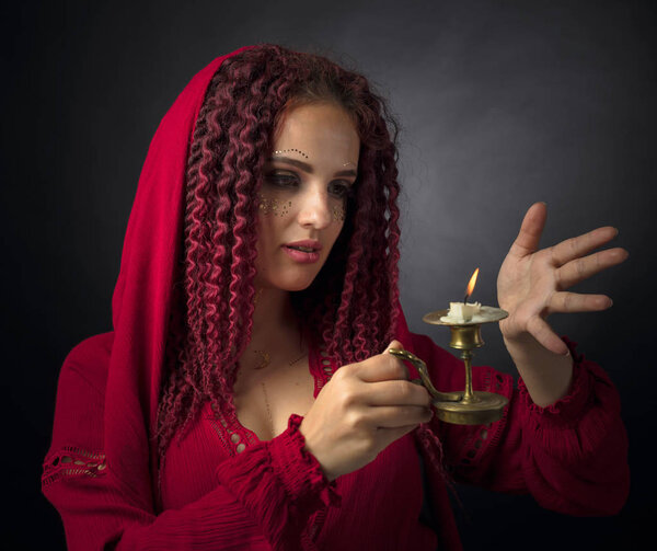 Portrait of attractive young woman in a fancy red dress with copper candlestick and burning candle. Girl with red wavy hair and makeup.