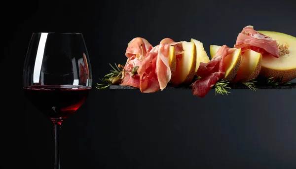 Glass of red wine and prosciutto with melon, capers and rosemary — Stock Photo, Image