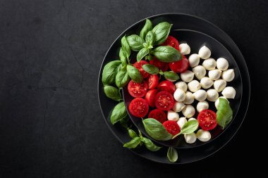 Domatesli Caprese salatası, mini mozzarella ve siyah tabakta fesleğen. İtalyan yemeği ve sağlıklı vejetaryen diyeti. Boşluğu kopyala, üst görünüm.