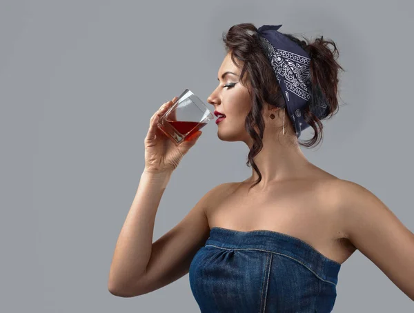 Beautiful young woman in a denim jumpsuit drinks whiskey. Portrait of a young brunette with wavy hair tied with a blue head kerchief. Copy space.