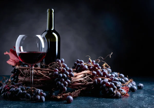 濃い青の背景に赤ワインと青ブドウのボトルとガラス 乾燥したブドウの葉は瓶の近くにある — ストック写真