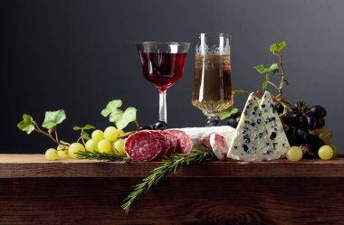 Eski ahşap bir masada kırmızı ve beyaz şarap ve atıştırmalıklar. Bir bardak şarap, mavi peynir, kurutulmuş sosis, üzüm ve biberiye. Basit ve lezzetli yemekler. Boşluğu kopyala.