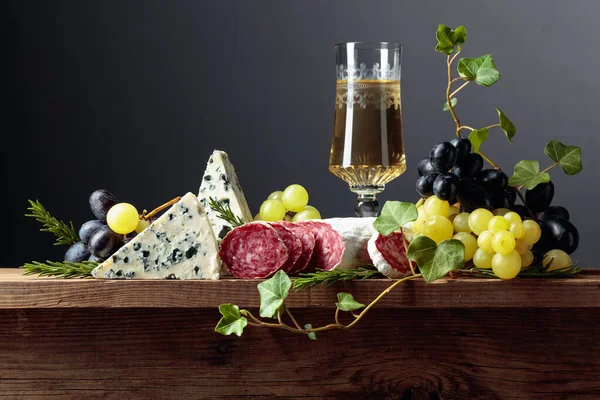 White wine and snacks on an old wooden table. Glass of wine, blue cheese, dry-cured sausage, grapes, and rosemary. Simple and tasty food. Copy space.