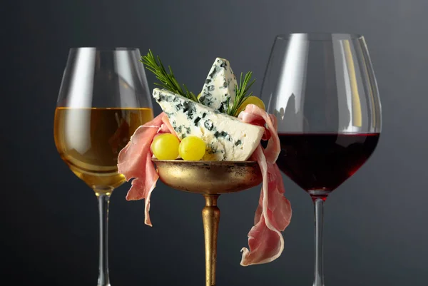 Red and white wine, blue cheese, prosciutto, grapes, and rosemary on a black background. Simple and tasty food. Copy space.