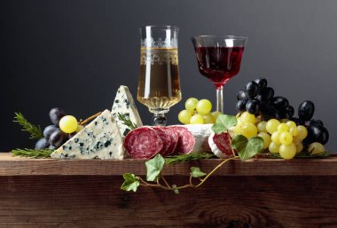 Şarap ve atıştırmalıklar eski ahşap bir masada. Bir bardak şarap, mavi peynir, kurutulmuş sosis, üzüm ve biberiye. Boşluğu kopyala.