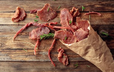 Eski ahşap bir masada, kağıt bir torbada biberiyeli baharatlı kurutulmuş et dilimleri. Üst görünüm.
