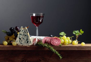 Eski ahşap bir masada kırmızı şarap ve atıştırmalıklar. Bir bardak şarap, mavi peynir, kurutulmuş sosis, üzüm ve biberiye. Basit ve lezzetli yemekler. Boşluğu kopyala.