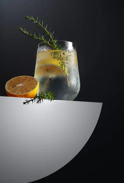 Alcohol drink (gin tonic cocktail) with lemon, juniper branch, and ice on a dark background, copy space. Iced cocktail drink with lemon.