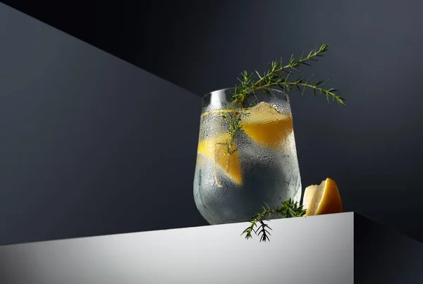 Alcohol drink (gin tonic cocktail) with lemon, juniper branch, and ice on a dark reflective background, copy space. Iced cocktail drink with lemon.