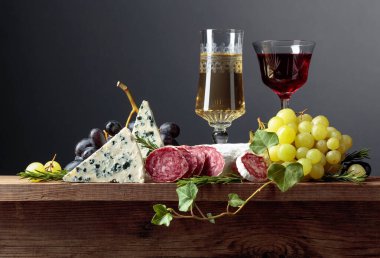 Eski ahşap bir masada kırmızı ve beyaz şarap ve atıştırmalıklar. Bir bardak şarap, mavi peynir, kurutulmuş sosis, üzüm ve biberiye. Basit ve lezzetli yemekler. Boşluğu kopyala.