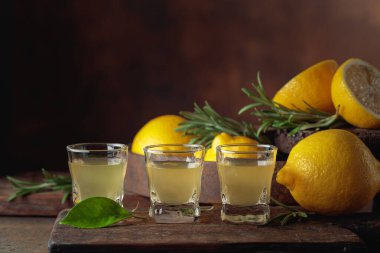 Kahverengi arka planda taze limon ve biberiye dallarıyla İtalyan likörü Limoncello..
