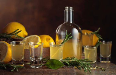 Kahverengi arka planda taze limon ve biberiye dallarıyla İtalyan likörü Limoncello..