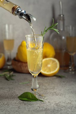 İtalyan likörü Limoncello şişeden bardağa dökülüyor. Taze limonlu ve gri taştan bir masada biberiye dallı tatlı alkollü içecek..