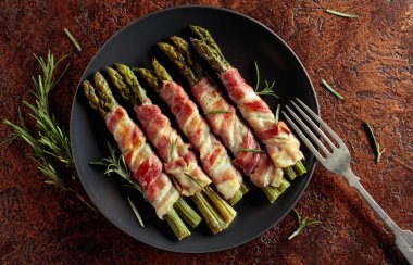 Domuz pastırmasına sarılı ızgara yeşil kuşkonmaz. Kahverengi arka planda siyah tabakta ev yapımı aperatif. Üst görünüm.