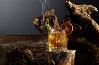 Eski usul bir kokteyl, buzlu, tahta bir kütükte. Kurutulmuş portakal dilimi ve yanan biberiye filizi ile süslenmiş soğuk, ferahlatıcı bir içecek. Boşluğu kopyala.