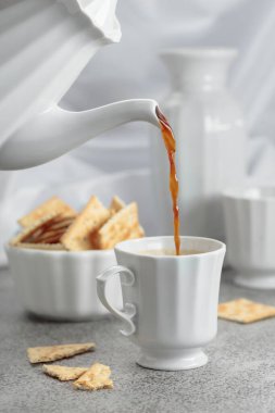 Beyaz porselen fincanda sade kahve ve gevrek kare kraker. Kahve demlikten fincana doldurulur. Gri taş masa ve beyaz çatal bıçaklar.