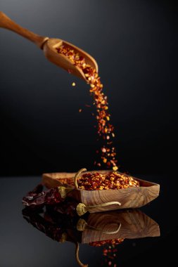 Kuru kırmızı biber ve siyah yansıtıcı arka planda ezilmiş kırmızı biber. Taneler ahşap bir kaseye dökülüyor..
