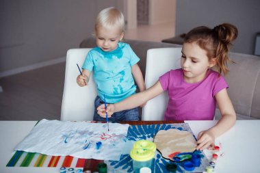 İki mutlu çocuk sarışın erkek ve boyalar ve fırçalar çizim ve masada evde oturan kız.