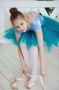 Pembe pointes ile mavi bir etekle genç balerin bir beyaz tahta bankta oturur.