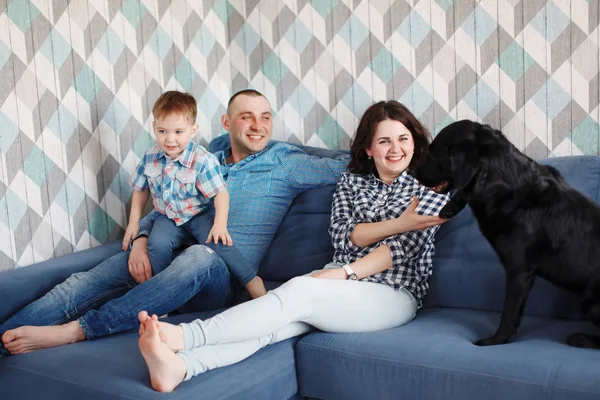 Young family mother, father and son sitting on a blue sofa with a black labrador retriever dog, playing and smiling