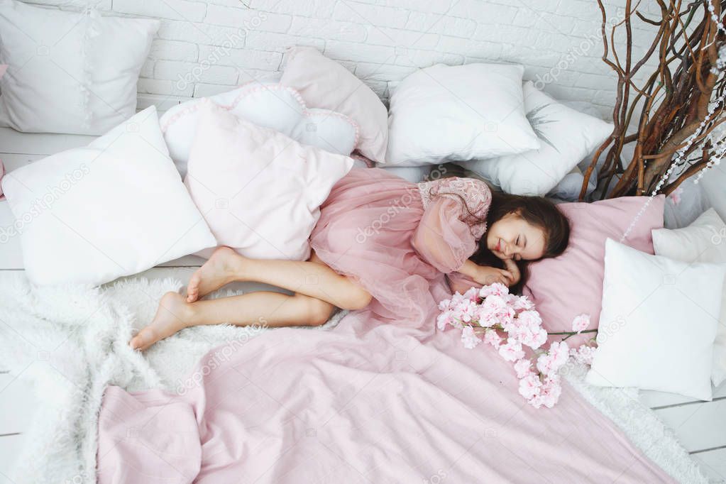 Little girl in a pink dress lies on a bed with white pillows and flowers