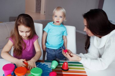 Genç bir hoş anne kızı ve oğlu ile. Sarışın çocuk ve kız ve kadın hamuru ile ve evde masada oturan pembe, kırmızı ve yeşil su bardağı ile oynama.