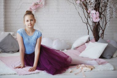 Genç balerin mavi giysiler ve mor etek pembe pointes ile beyaz bir yatak yastık ile oturur.