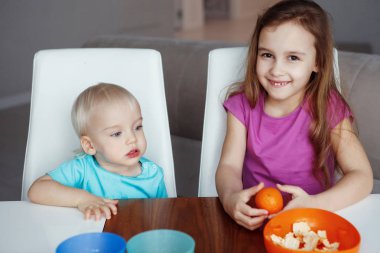 İki mutlu çocuk evde oturan oynayan sarışın erkek ve kız yiyor portakal, Masa