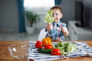 Sevimli küçük sarışın çocuk masada yeşil, sarı ve kırmızı sebze salata yapmak hazırlık yapıyordu