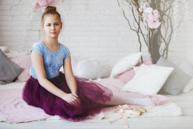 Genç balerin mavi giysiler ve mor etek pembe pointes ile beyaz bir yatak yastık ile oturur.