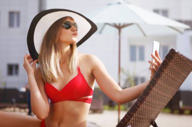 Genç kadın bir kırmızı mayo güzel ve mavi su resort otel ile yapay rattan havuz kenarında şezlong içinde oturan smartphone içinde plaj şapka görünüyor.