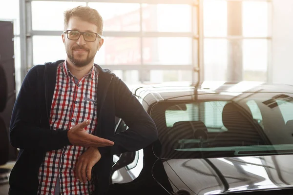 Young guy in a plaid shirt and glasses is standing in the car dealership and pointing at the car that he has just bought. The concept of buying vehicle.