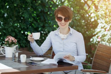 Yeşil yaprakları bir arka plan karşı gülümseyen güneş gözlüğü içecekler kahve beyaz bir fincan bir yaz kafede Genç Sevimli Kadın.