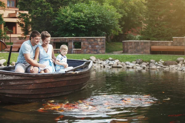 Mother, father and son sit in a wooden boat with oars and feed the fish with bread.