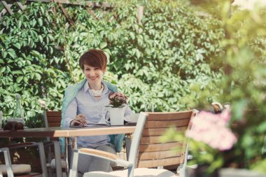 Bakarak bir smartphone bir yaz kafede yeşil yaprakları bir arka plan karşı gülümseyen bir beyaz fincan genç Sevimli Kadın içecekler kahve.