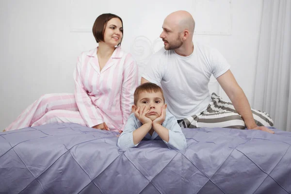Young beauty and bald bearded man in pajamas look at each other in bed with blue bedclothes behind her son.