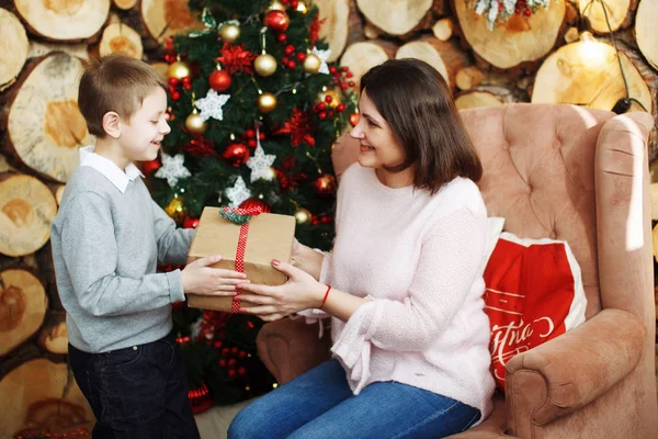 The boy gives his mother a gift in a box near the Christmas tree.