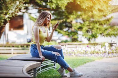 Genç oldukça şık kız sarı bir tişört ve kot bir smartphone vasıl seyir ve kentsel bir ortamda gülümseyen.