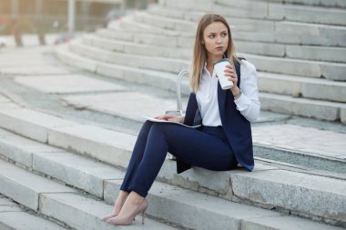 Bir deste kağıt ve bir karton kahve Kupası'na bir koyu mavi takım elbise ve beyaz gömlek ile oldukça neşeli kadın yaz şehirde taş basamaklar üzerinde oturuyor.
