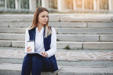 Bir deste kağıt ve bir karton kahve Kupası'na bir koyu mavi takım elbise ve beyaz gömlek ile oldukça neşeli kadın yaz şehirde taş basamaklar üzerinde oturuyor.