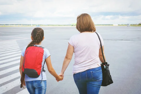 Mom with a bag and a daughter with a red backpack passengers go to the ramp of the plane on the airport field.