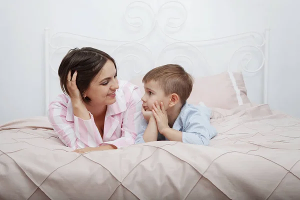 Young, pretty woman and son in pink and blue pajamas in bed with bedding and pillows hug and laugh.