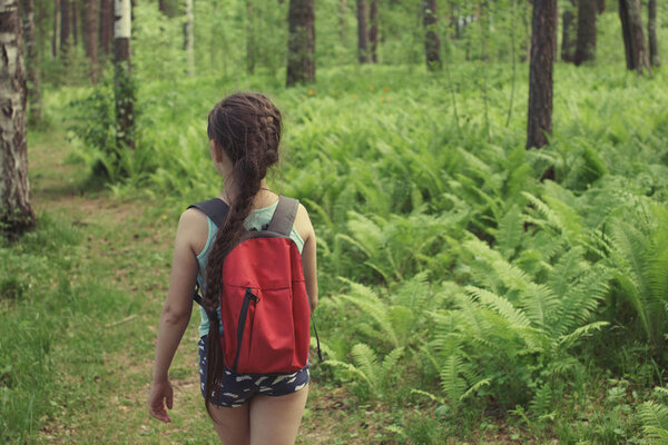 Teenager girl with a pigtail, in blue shorts, with a red backpack is traveling along a path in a green summer forest.