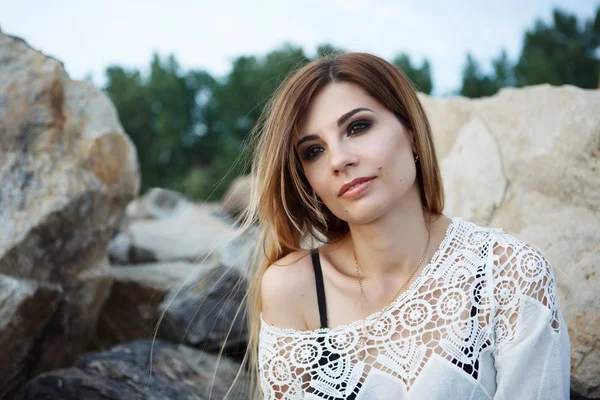 Young beautiful woman in a white tunic sits on gray granite stones.