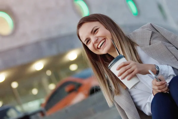 Pretty cheerful woman smiling with paper coffee cup in a grey jacket and white shirt is sitting on the stone steps in the summer city.