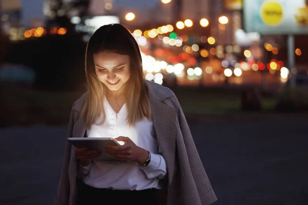 美しい若い女性は マルチカラーのライトの背景に夕方の都市の路上でタブレットを見ています デバイスの光に顔を照らされました — ストック写真