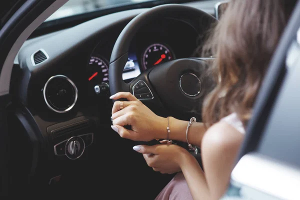 Nice beautiful girl with long curly hair is sitting and holds the steering wheel in a black car. Just buy a car in the dealership. The concept of buying a vehicle.